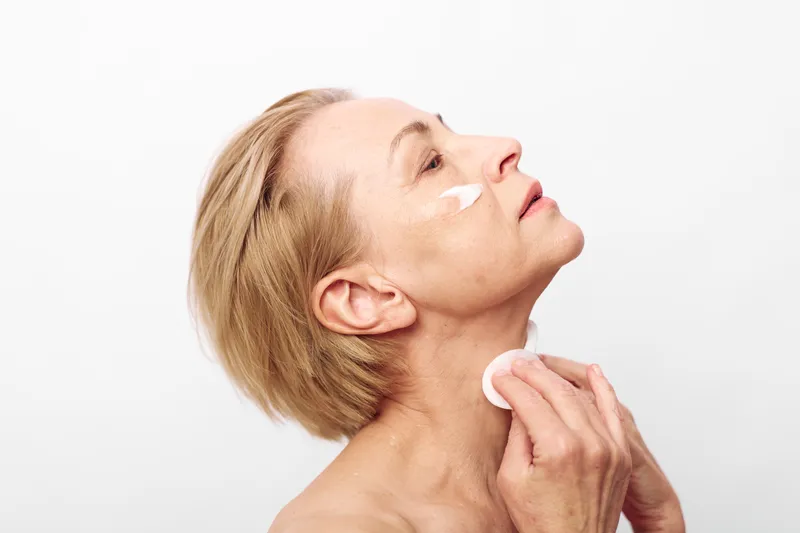 Mature woman applies facial cream and uses cotton pad on neck, showing skincare routine and self-care for aging skin in bright studio with white background and natural light.