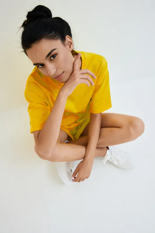 Young woman in bright yellow casual outfit sitting on white floor, looking thoughtfully with hand near face in minimalist studio setting.