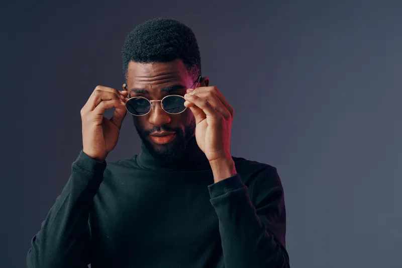 Stylish young Black man adjusting sunglasses against a dark gray background, expressing confidence and modern fashion with a sleek look