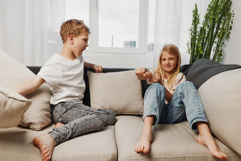 Playful young boy and girl sitting on beige sofa in bright living room having fun and interacting, casual clothing and relaxed atmosphere. Concept for childhood, friendship, and home life.
