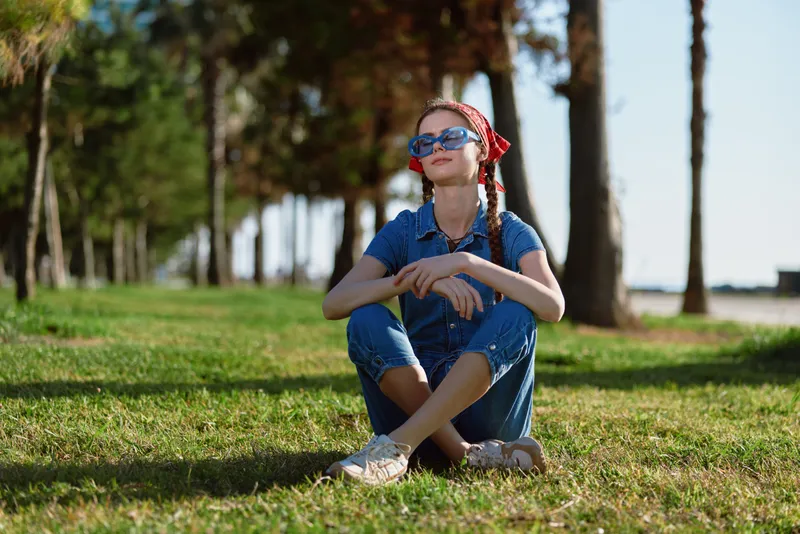 Relaxed woman sitting on grass wearing sunglasses with braided hair and casual denim outfit in bright sunny park setting during daytime.