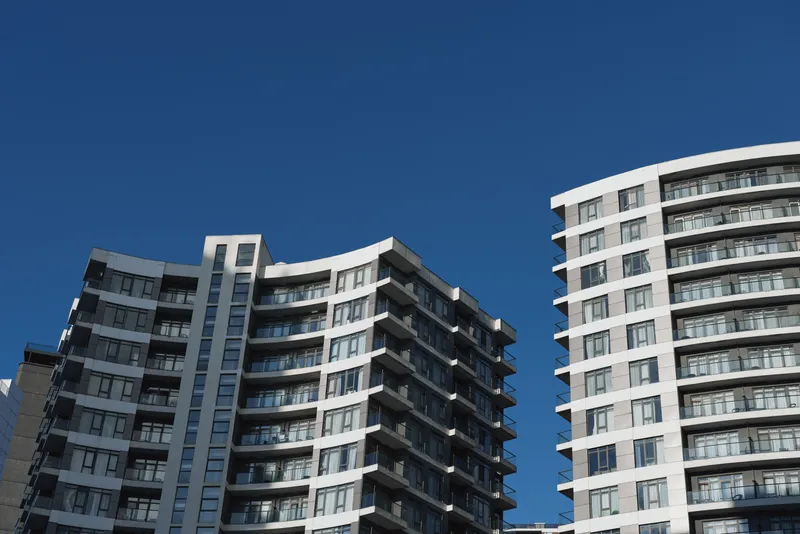 Modern high rise apartment buildings rise against a clear blue sky, highlighting angular balconies, repeating geometric patterns, and a sleek urban living aesthetic.