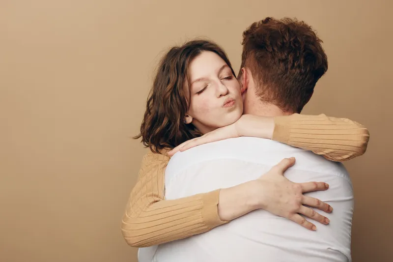 Young woman with brown hair hugs man wearing white shirt, eyes closed and lips puckered. Intimate moment of affection and love on beige background in casual clothing.