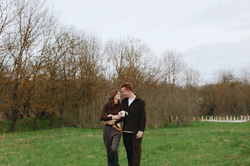 Cheerful couple outdoors with a playful goat kid, embracing in a green field, soft breeze, natural colors, warm interaction, candid moment, rustic backdrop, everyday romance, joyful connection,