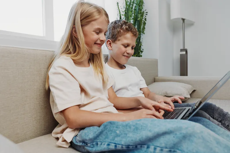 Two happy children sitting on sofa using laptop together in cozy living room. Concept for learning, technology, childhood, and family leisure at home environment.