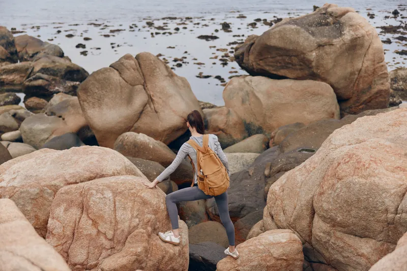 Young woman exploring rocky coast with a backpack, wearing athletic clothing, showcasing adventure and nature in a serene environment