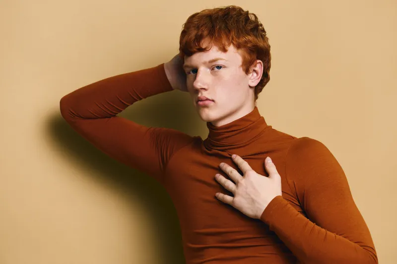 Portrait of young man with red hair and freckles wearing brown turtleneck posing with hand on head against beige background. Fashion, style and confident look in studio.