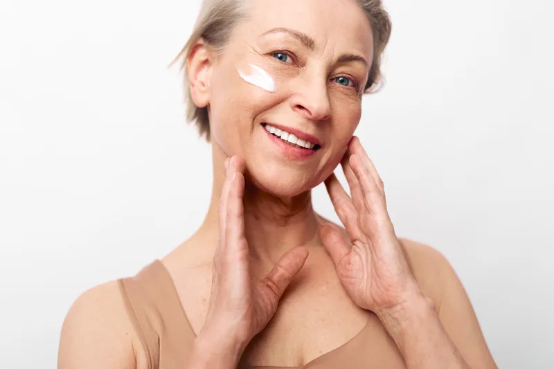 Smiling mature woman with natural skin applies white face cream on cheek. Close-up on light background. Concept of skincare, antiaging, beauty and selfcare for older adults.