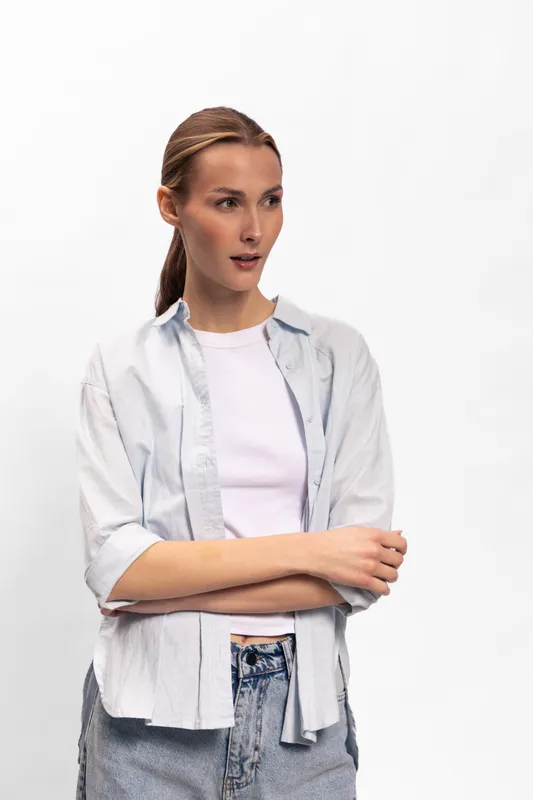 Young woman with long hair tied back wears casual shirt and jeans, standing with arms crossed against white background. Concept of casual style, confidence, and thoughtful expression.