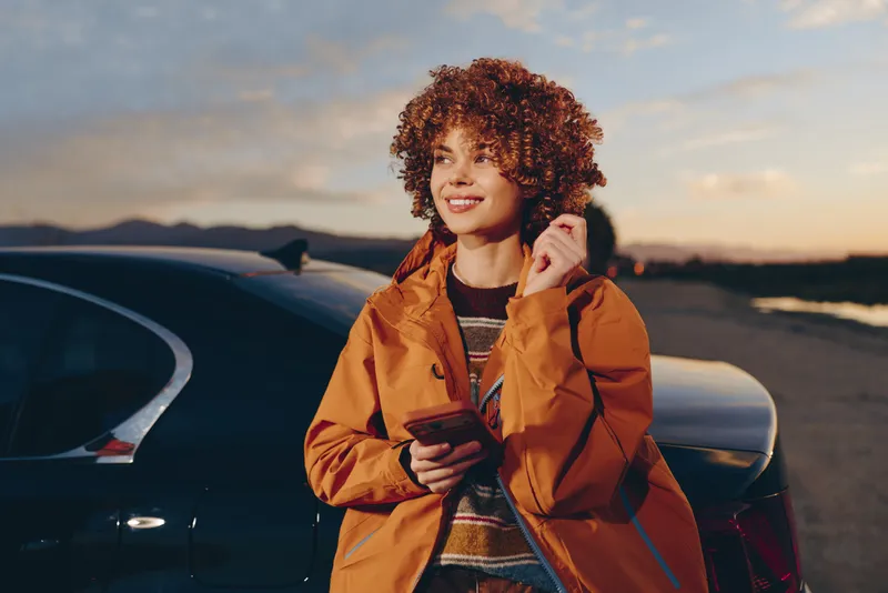 Smiling woman lifestyle outdoors near a parked car at sunset, curly hair, orange jacket, holding a smartphone, relaxed stance, warm evening light, coastal road background, soft breeze