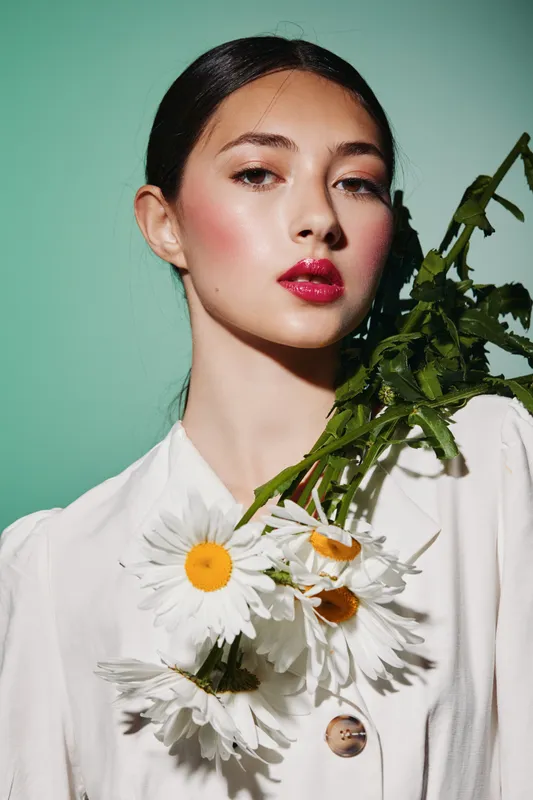 Young woman with bold red makeup and sleek hair holds white daisies wearing a stylish white blouse. Editorial fashion shot on green background for beauty concepts.