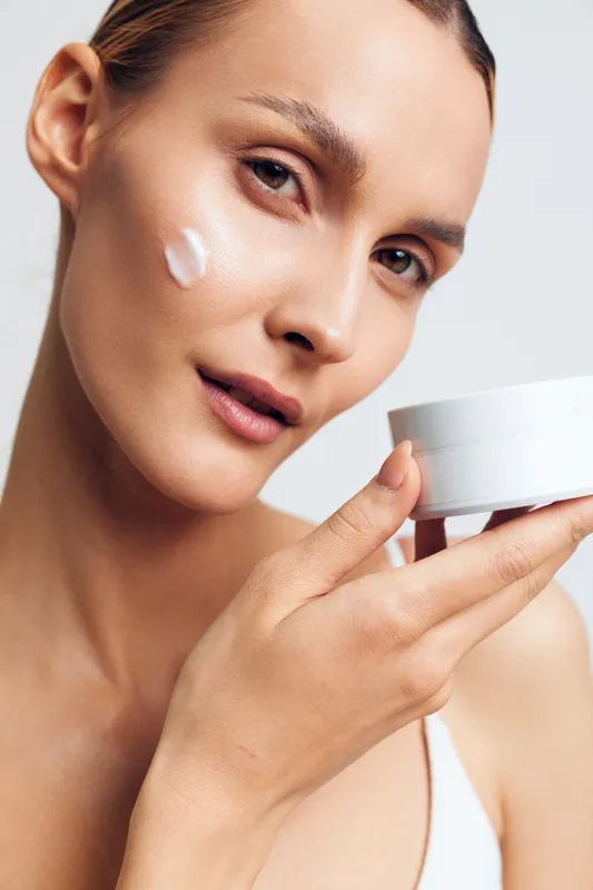 A young woman with clear skin applies white cream on her cheek while holding an open jar. The close-up shows skincare routine and natural beauty concept with soft lighting.
