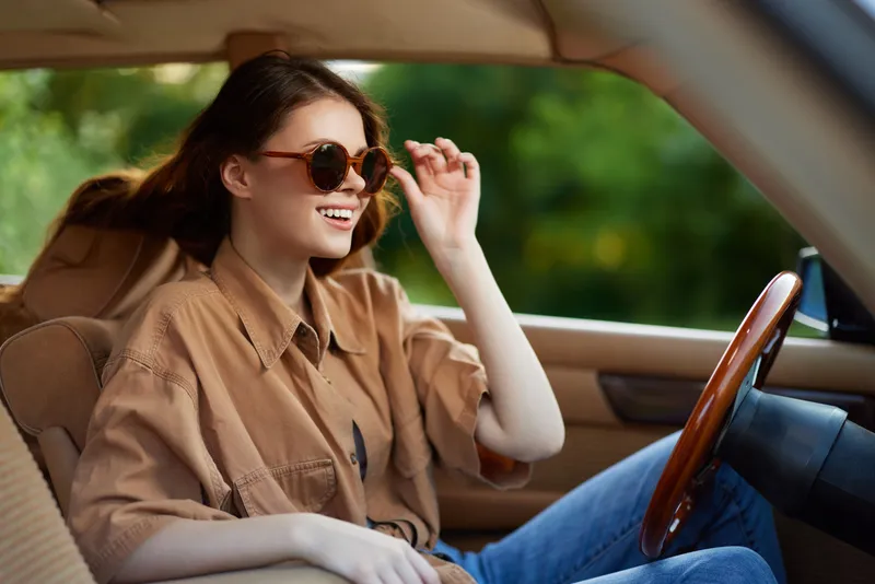 Young woman enjoying a road trip in a convertible car, wearing sunglasses and a casual outfit, with a joyful expression and a vibrant outdoor background.