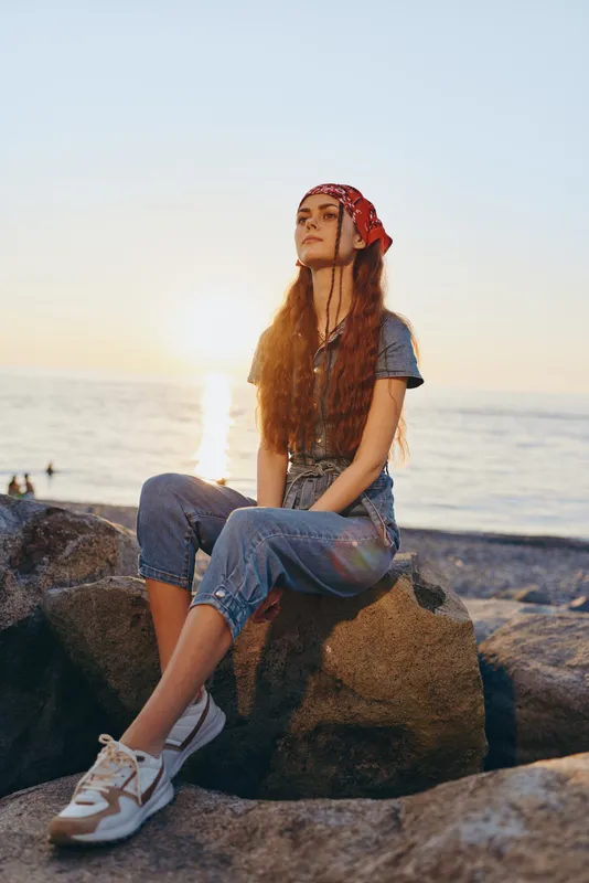 Young woman with long red hair sitting on rocks by the sea during golden hour wearing a red bandana, casual clothes, and white sneakers, relaxed and thoughtful mood