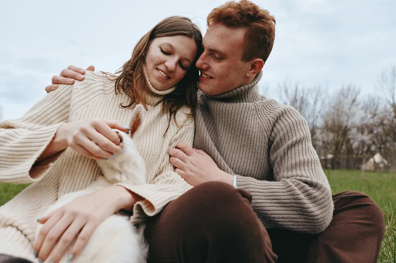 Smiling couple in chunky sweaters outdoors, caressing a fluffy white dog, capturing a cozy moment of love and companionship. Natural light, soft shadows, rural backdrop, relaxed mood, wellness and