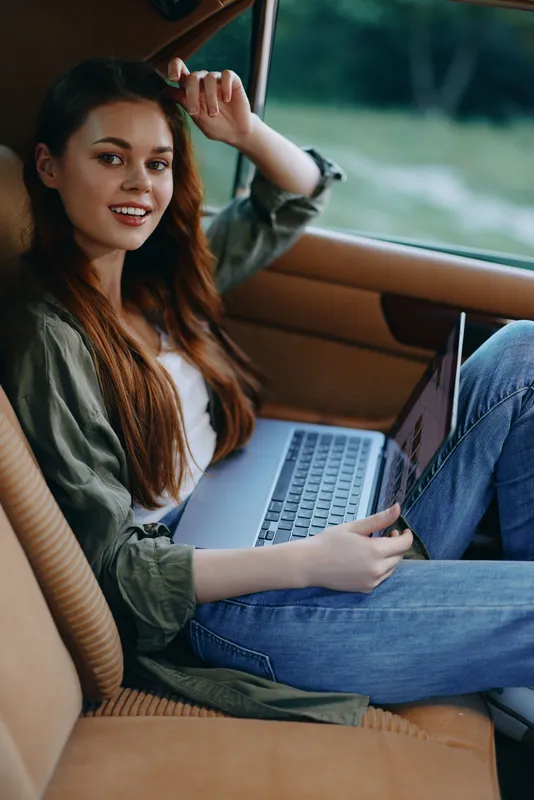 Young woman smiling while working on a laptop in a car, dressed casually in jeans and a green jacket, enjoying the moment in a natural outdoor setting.