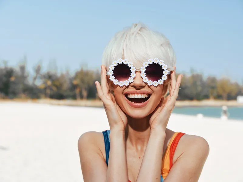 Bright portrait of a joyful woman on a sunny beach, wearing round flower sunglasses and a colorful swimsuit, capturing a playful summer moment with warm sunlight, sparkling water, and a radiant smile.