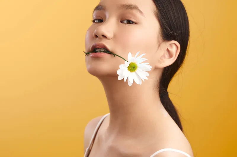 Asian woman with a flower between lips stands against a warm yellow backdrop, exuding playful poise and modern elegance in a minimalist studio setting.