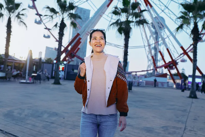 Smiling woman in a cozy jacket strolls along a palm lined park boardwalk, enjoying sunny outdoor ambiance and urban leisure, capturing a candid moment of happiness and exploration