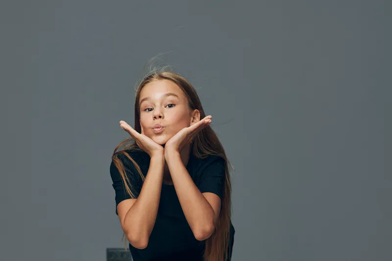 Girl posing with hands on cheeks making a playful kiss face with long hair and casual t shirt on plain gray background. Child, expression and youth with fun gesture in studio.