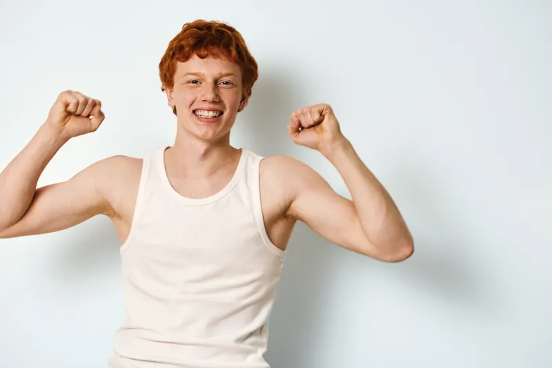Young man with red hair in white tank top smiling and raising fists showing strength and happiness standing against plain light background with energetic posture and expression
