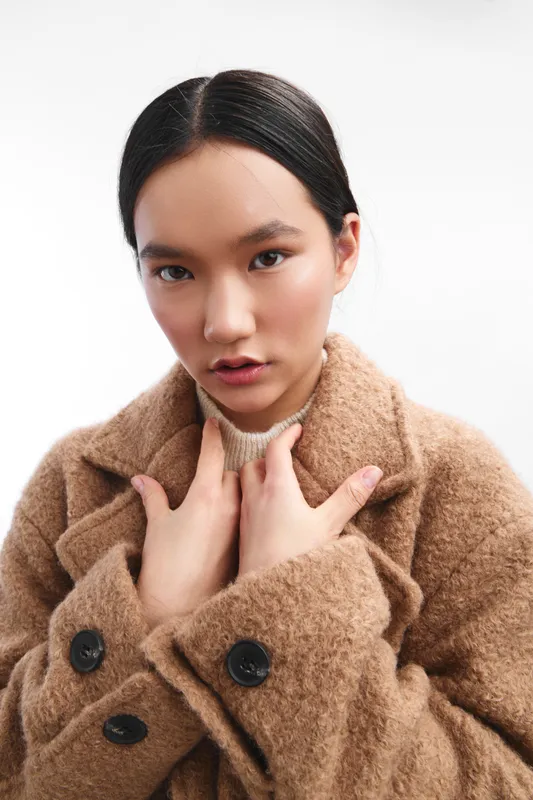 Asian woman wearing a warm brown coat sits against a clean backdrop, offering a stylish, intimate portrait that emphasizes texture, mood, and understated elegance.
