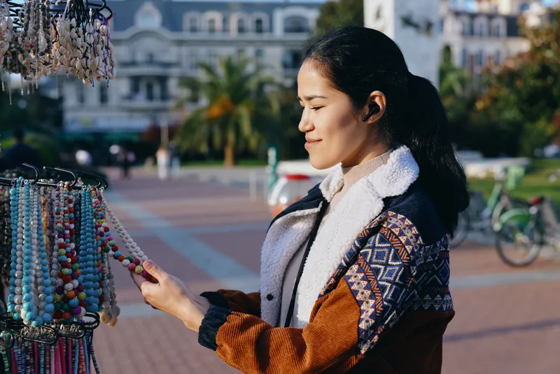 Jewelry beads on display at an outdoor market, a shopper examines colorful strands, showcasing handmade accessory craft and a sunny street atmosphere