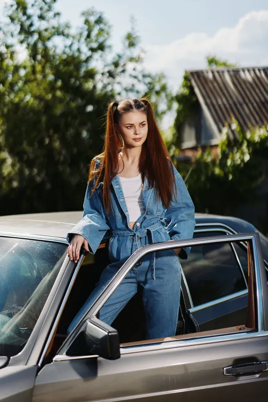 Young woman in denim outfit poses confidently on vintage car, enjoying sunny day outdoors, showcasing casual fashion and leisure vibes.