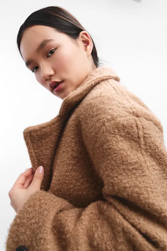 asian woman wearing a warm brown coat poses in a clean studio, soft lighting highlights texture and her thoughtful expression, creating a refined fashion portrait.