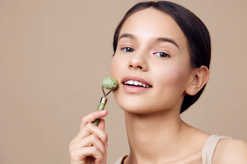 Young woman using a jade roller for skincare on a neutral background, promoting healthy skin and self care rituals with a fresh, glowing appearance