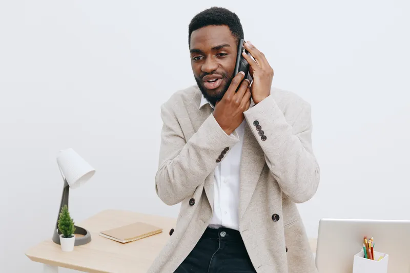 Professional young African man in casual blazer smiling and talking on mobile phone while standing at a modern office workspace with white background, desk lamp, and potted plant. People lifestyle con