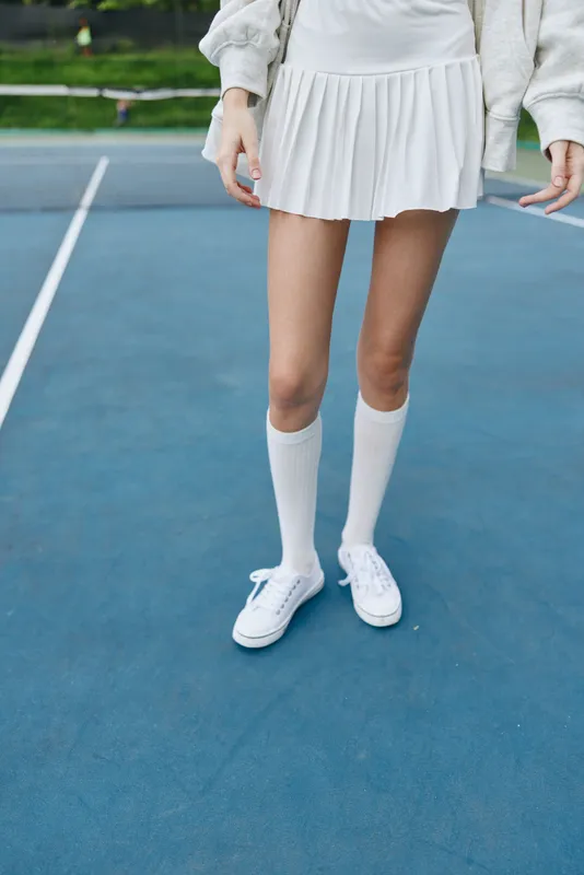Young woman in a white tennis skirt with knee high socks and sneakers on a blue tennis court, exuding a sporty and energetic vibe during a sunny day