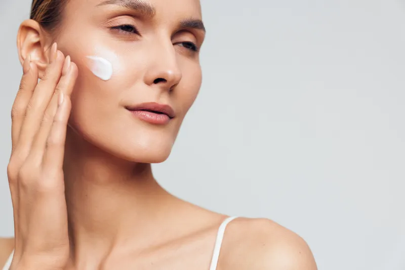 Close-up of a young woman applying white cream to her cheek, with a soft neutral background, capturing skincare routine, wellness, and self-care in a calm intimate moment highlighting skin texture
