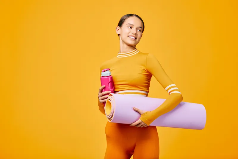 Fitness, woman, sport and workout scene with a confident female in orange activewear holding a water bottle and yoga mat against a vibrant orange backdrop.