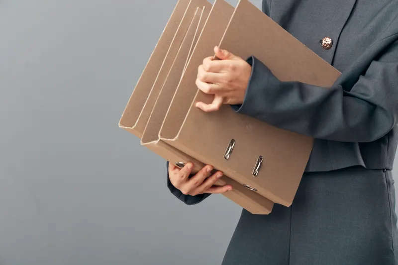 Businesswoman holding multiple large brown file folders with metal rings on gray background. Office worker in dark suit carrying important documents with hands and professional attire.