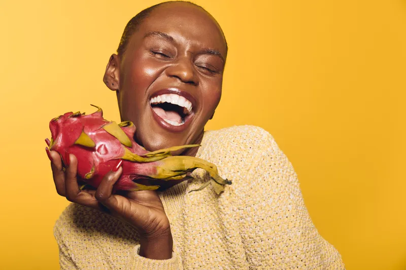 Happy black woman with dark skin laughing and holding a dragon fruit near her face on yellow background. Concept of healthy lifestyle, fresh fruit, and natural beauty.