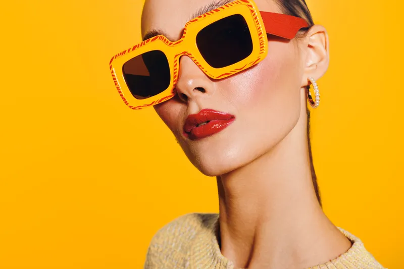 Stylish woman with bright orange sunglasses and red lipstick poses against vibrant yellow background. Fashion portrait with modern accessories and confident expression.