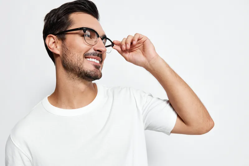 Smiling man wearing eyeglasses and casual white t-shirt, looking cheerful while touching glasses, set against a plain light background. Lifestyle concept.