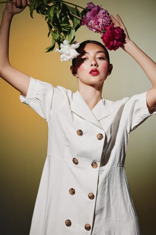 Fashion model wearing stylish white dress poses holding colorful flowers above her head. Bold red lipstick, natural makeup, creative beauty studio photo with warm lighting. Fashion, beauty, creative.