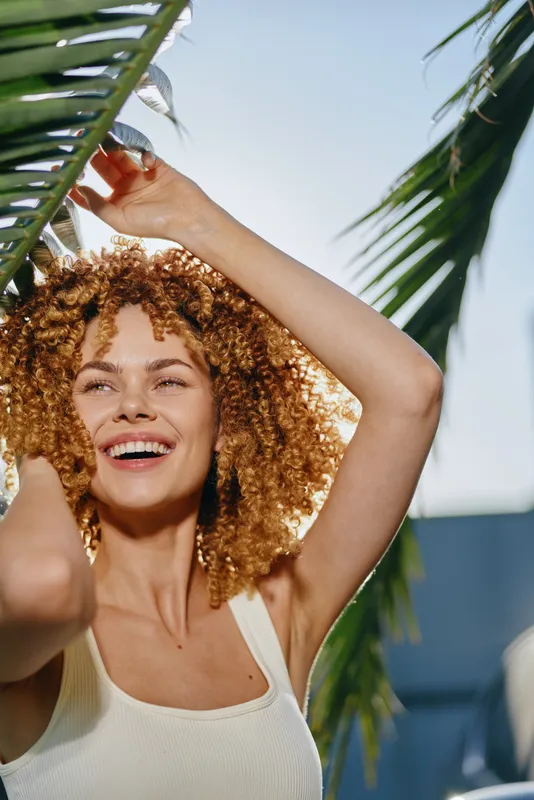 smiling woman with curly hair enjoys sunny day outdoors under green palm leaves, wearing white tank top, natural light, fresh summer atmosphere