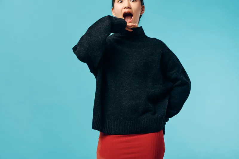 Young woman with surprised expression wearing black sweater and red skirt standing against blue background. Casual clothing, fashion, emotion, reaction and studio portrait in lifestyle concept.