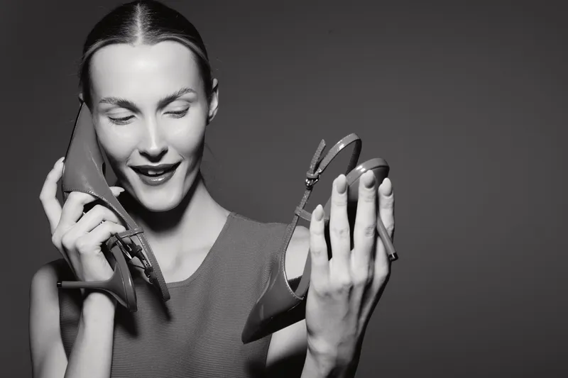 Young white woman with pale skin holds pair of high heels near face smiling, isolated on dark background. Concept of fashion and style.