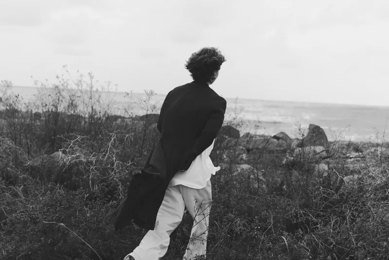Running silhouette along a windy coast, a figure in a coat heads toward the sea, black and white mood with sparse dunes and rugged rocks, conveying motion, freedom, and coastal solitude.