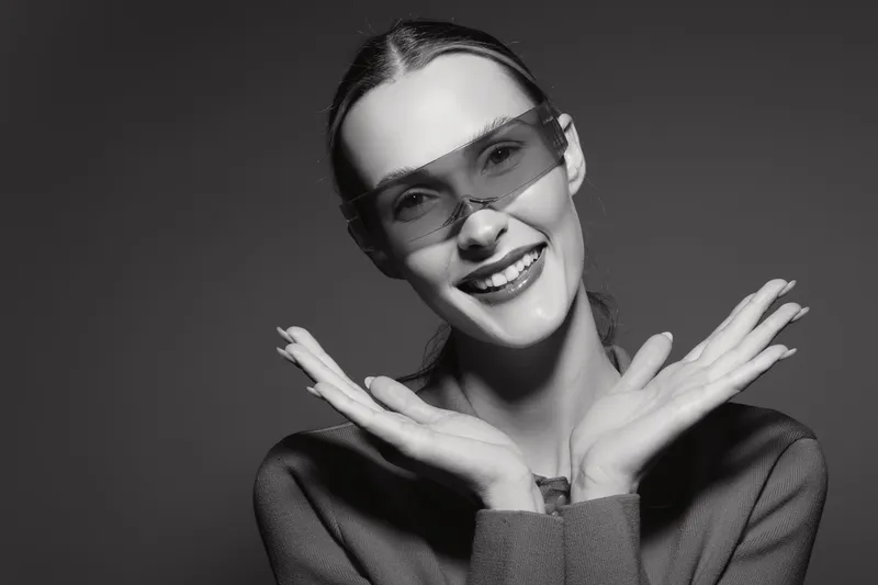 Smiling caucasian woman with fair skin wearing big sunglasses and a sweater poses happily against a dark background in studio lighting.