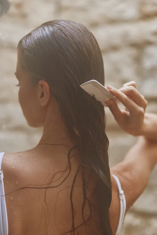 Woman combing wet long hair outdoors with white comb, natural light, back view, beauty and hair care concept, summer mood, soft beige stone background.