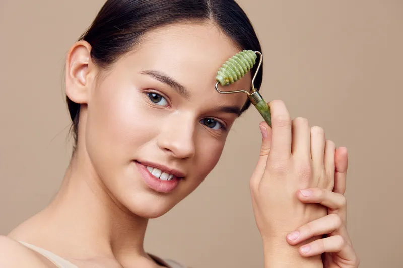 Young woman using jade roller on her face for skincare routine, promoting healthy skin and beauty concepts with serene expression and neutral background