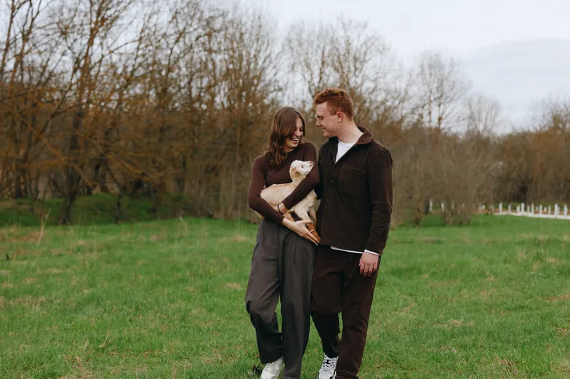 Smiling couple walking through a green field with a curious goat kid, sharing a warm lifestyle moment outdoors. Casual outfits, soft daylight, and playful companionship convey relaxed, wholesome