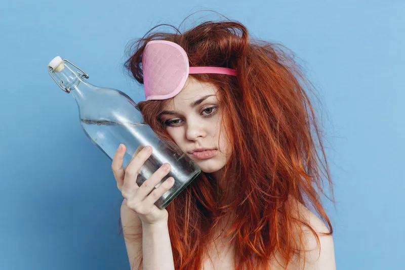 Эмоциональная рыжеволосая женщина с маской для сна, похмелье, в руках бутылка алкоголя