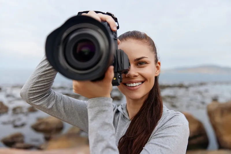 Young woman with a camera by the seaside, showcasing joy and passion for photography in sporty attire, surrounded by natural beauty and soft colors