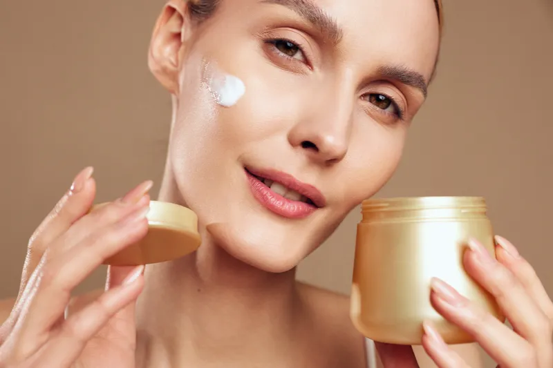 Close up of woman applying white face cream with her hand while holding a skincare jar on warm beige background Soft studio lighting shows self care wellness beauty and spa routine